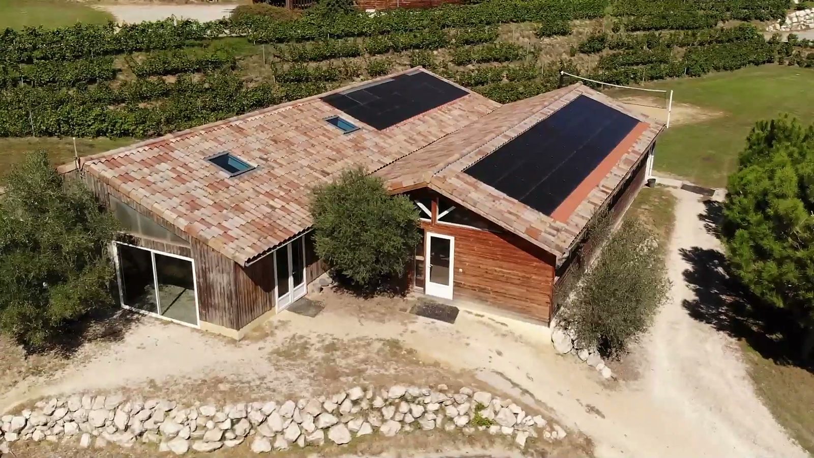 Vu du ciel, un bâtiment du domaine en bois et tuiles, son toit couvert de panneaux solaires, au bord de la vigne