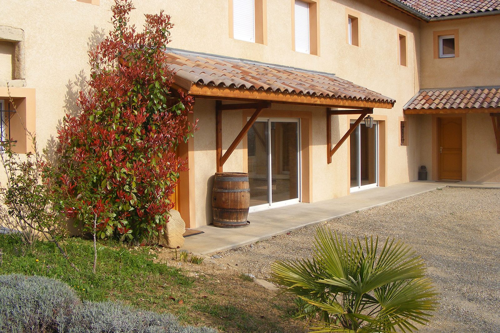 La façade du Marronnier, son auvent en tuiles, un tonneau et un photinia rouge